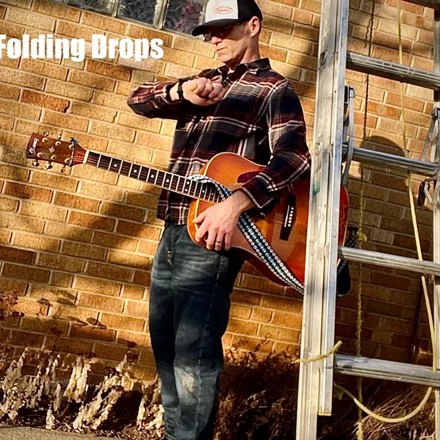 Jim Crowther of Folding Drops holding an acoustic guitar in front of a brick wall